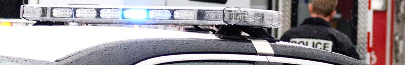 Image of the top of police car with emergency light bar on top. In the background is a police officer whose back is facing the camera with the words POLICE across the back of his uniform. The officer is facing a fire truck.